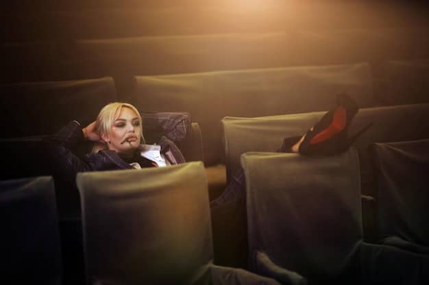 A woman is sitting alone in a dim movie theatre, staring intently at the screen with a mix of wonder and curiosity. The scene is lit primarily by the light from the movie screen.