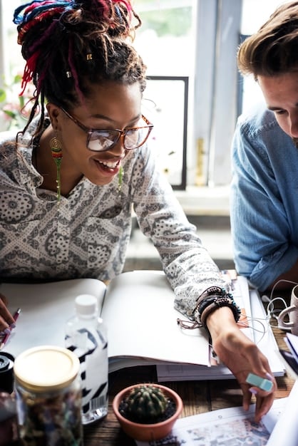 Two people from different cultural backgrounds working together on a creative project. They are engaged in a deep conversation, surrounded by books and art samples that represent their respective cultures, symbolizing a collaborative and respectful creative process.